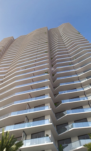 Tall modern apartment building with numerous balconies, viewed from below against a clear blue sky. The structure appears sleek and elegant.