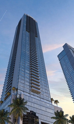 Tall, glass skyscraper reflecting the sunset sky, surrounded by palm trees. A second tower is visible in the background. Modern and serene atmosphere.