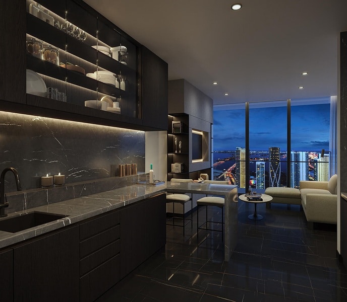 Modern kitchen and living room at night, featuring dark cabinets, a marble countertop, under-cabinet lighting, and a cityscape view through large windows.