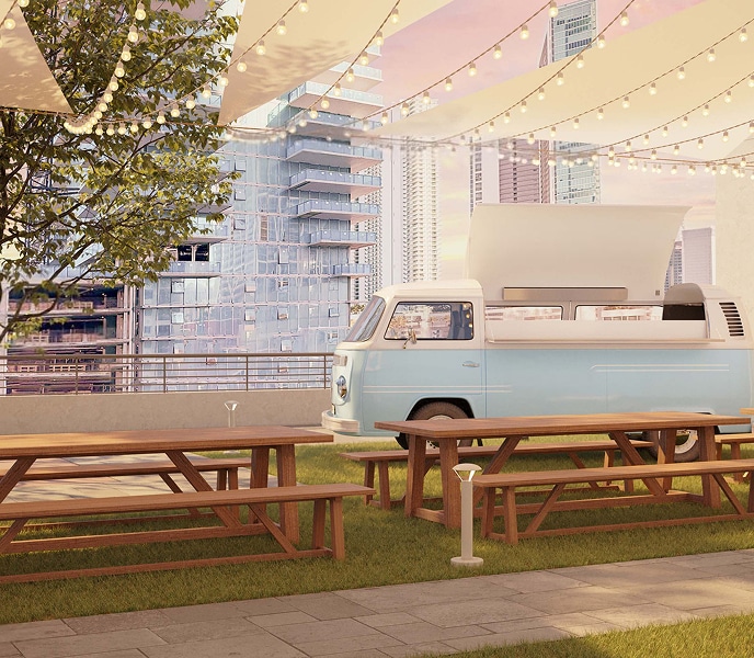 A pastel blue vintage van serves as a food truck amidst picnic tables on grass. Overhead string lights create a cozy ambiance against a city skyline at sunset.