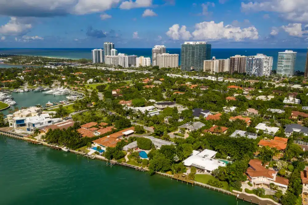 Aerial view of Bal Harbour bayfront homes with private docks, lush tree-lined streets, luxury residences, and oceanfront towers in the distance.