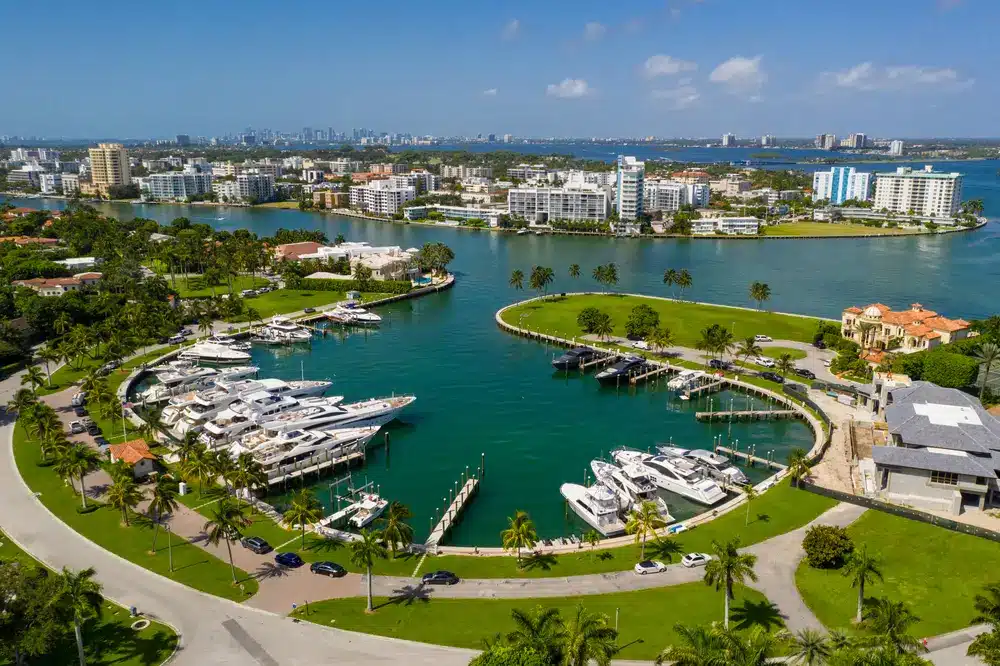 Aerial view of Bal Harbour marina with luxury yachts, calm bay waters, palm-lined shoreline, and waterfront residences in the background.