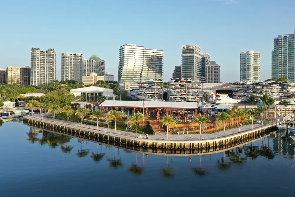 Waterfront marina in Coconut Grove with palm-lined promenade, docked yachts, waterfront dining, and high-rise buildings in the background.