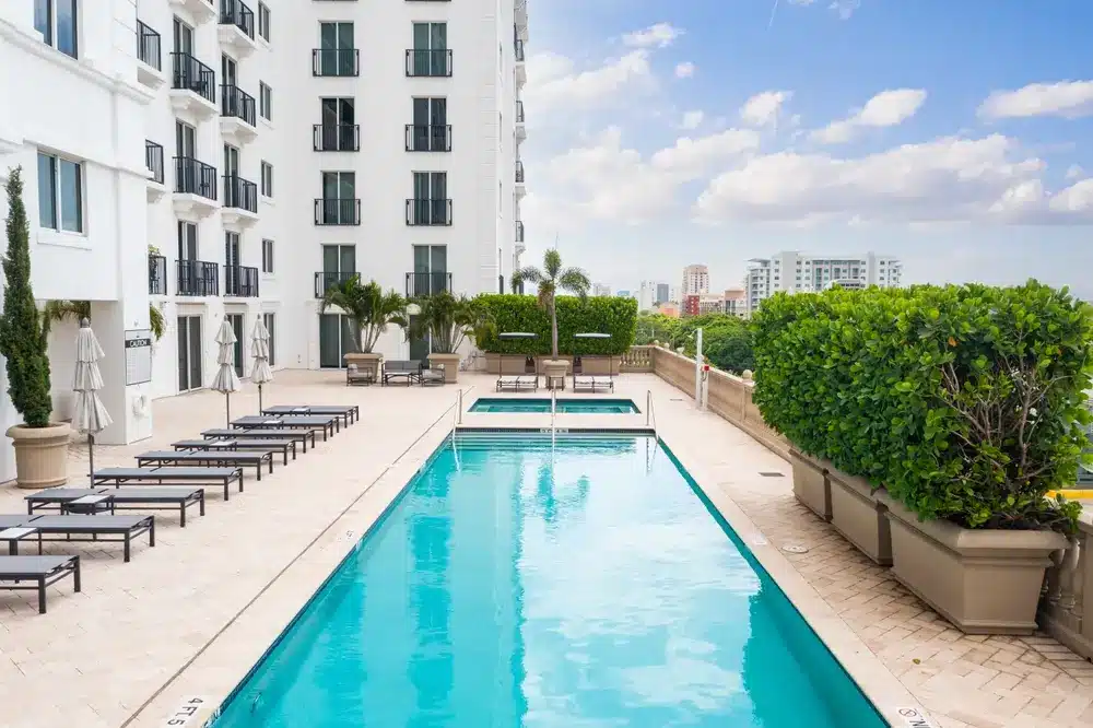 Condominium pool deck in Coral Gables with lounge seating, landscaped planters, modern architecture, and city views under a clear sky.