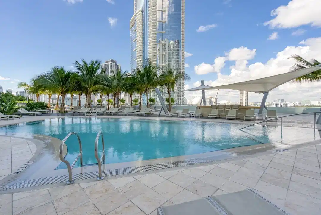 Rooftop swimming pool in Downtown Miami with palm-lined sundeck, modern lounge seating, shade structures, and high-rise glass towers in the background under a clear blue sky.