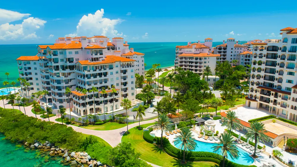 Aerial view of luxury condominiums on Fisher Island with Mediterranean-style architecture, palm trees, resort pools, and the Atlantic Ocean beyond.