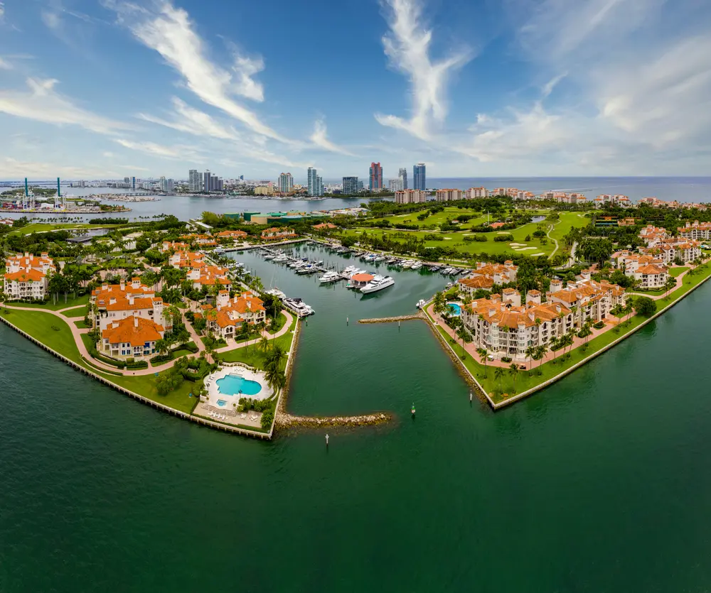 Aerial view of Fisher Island with luxury waterfront residences, private marina, manicured golf greens, and Biscayne Bay with Miami skyline beyond.