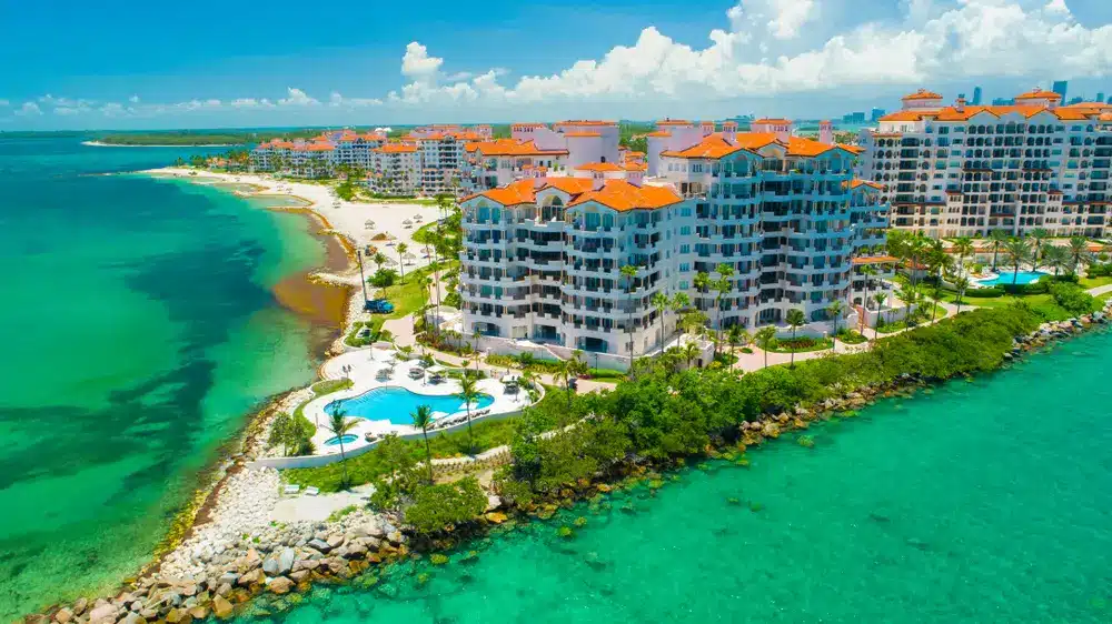 Aerial view of oceanfront condominiums on Fisher Island with palm-lined grounds, resort-style pools, sandy beach, and turquoise waters.