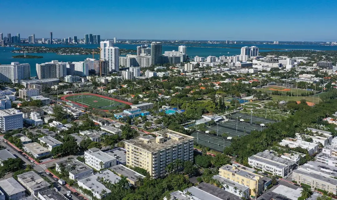 Aerial view of Flamingo Park in Miami Beach showing residential streets, tennis courts, athletic fields, surrounding low-rise buildings, and Biscayne Bay with the Miami skyline in the distance.