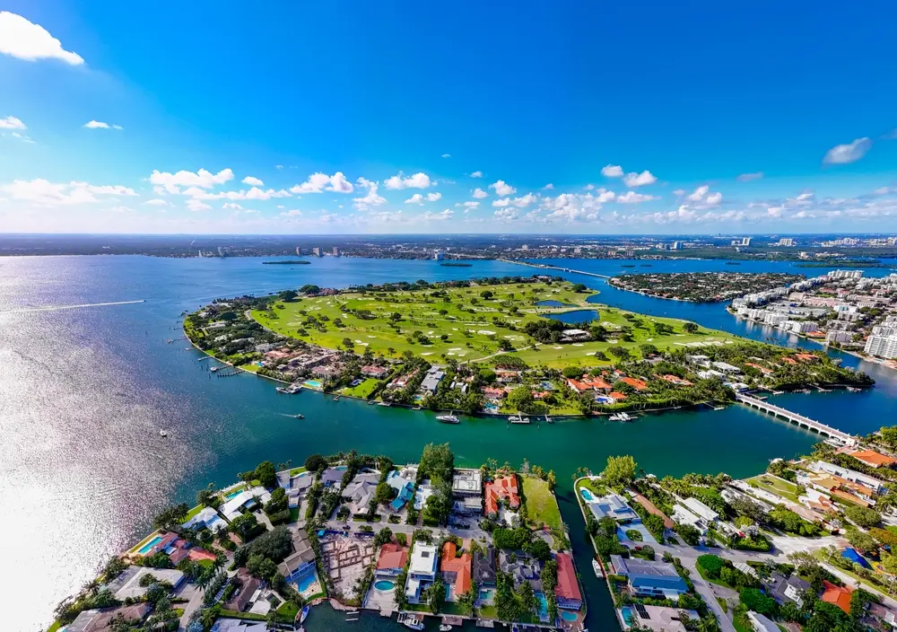 Aerial view of Indian Creek Island with private waterfront estates, central golf course, Biscayne Bay, and surrounding luxury communities.
