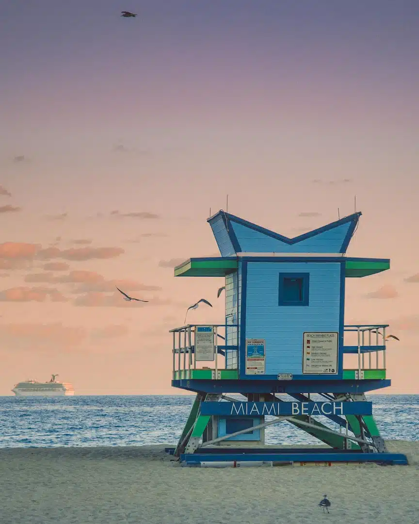 Miami Beach lifeguard tower at sunset with pastel sky, flying seabirds, calm ocean waters, and cruise ship on the horizon.