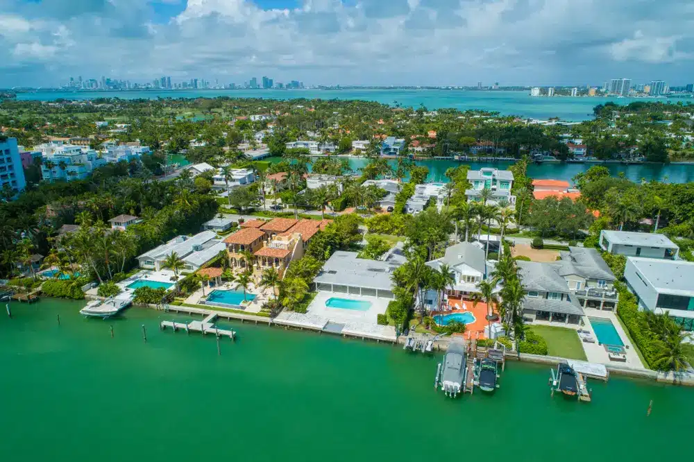 Aerial view of waterfront homes in Miami with private docks, pools, palm trees, and calm bay waters, with the city skyline in the distance.