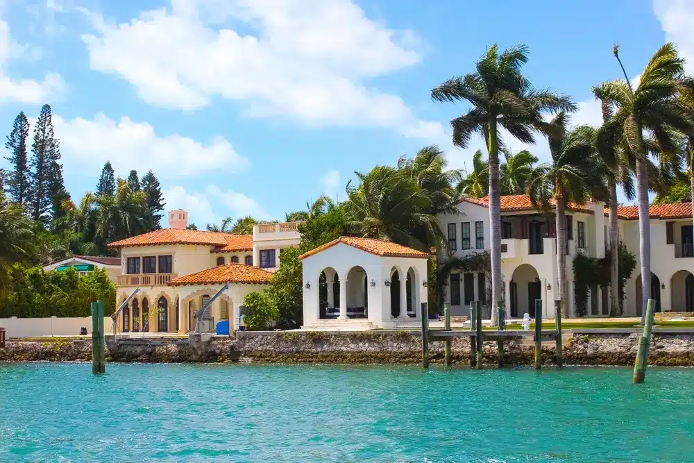 Waterfront estate in Miami Beach with Mediterranean-style architecture, palm trees, private dock, and calm bay waters under clear skies.