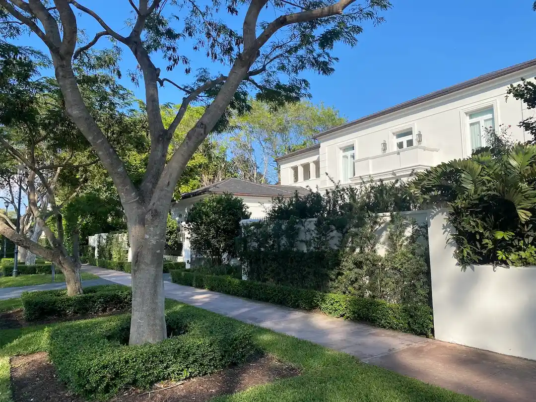 Luxury waterfront home along Pinetree Drive in Miami Beach with manicured hedges, gated entrance, and tree-lined residential sidewalk.