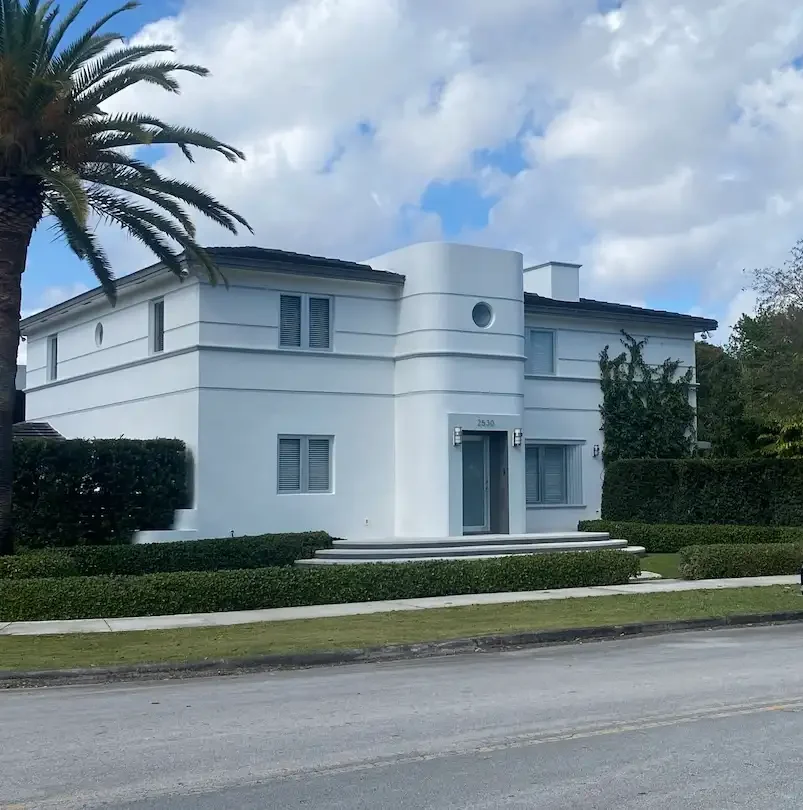 Modern two-story white residence on a quiet residential street in The Roads Miami, featuring clean lines, manicured hedges, and classic architecture.
